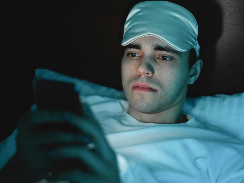 A person using a smartphone in bed at night, blue light glowing on their face, illustrating sleep disruption.
