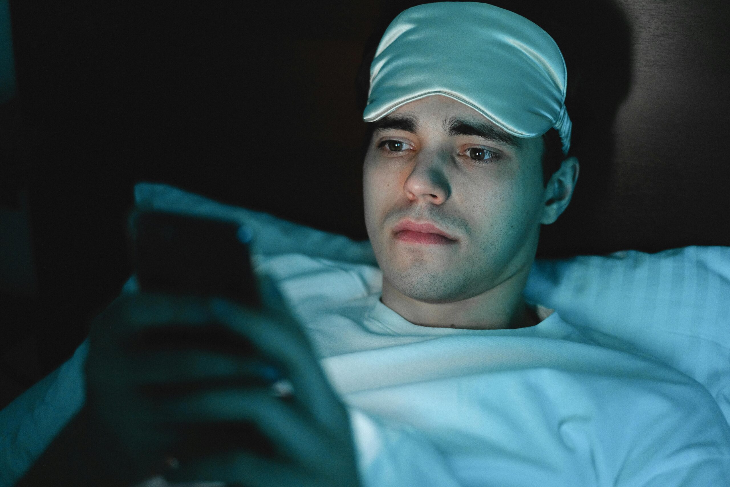 A person using a smartphone in bed at night, blue light glowing on their face, illustrating sleep disruption.