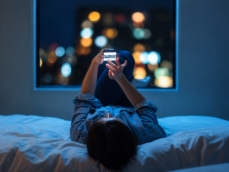 A person using a smartphone in bed at night, blue light glowing on their face, illustrating sleep disruption.